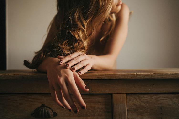 Crop unrecognizable female with wavy hair on face and manicure leaning with hands on wooden table in house