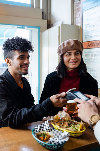 Cheerful young ethnic couple paying for food order with smartphone while standing near counter in light restaurant