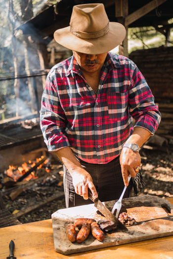 Argentinian male in hat with knife and carving fork cutting yummy meat piece on board against barbecue rack in sunlight
