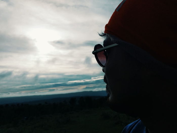 Close-up of man wearing sunglasses against cloudy sky during sunset