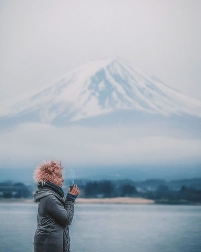 Side view of young woman having drink while standing by lake during winter