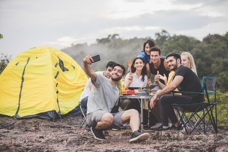 Friends taking selfie while sitting on land by tent against sky
