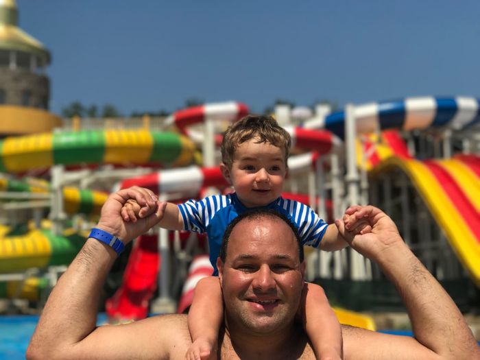 Portrait of father carrying son on shoulders at water park during sunny day