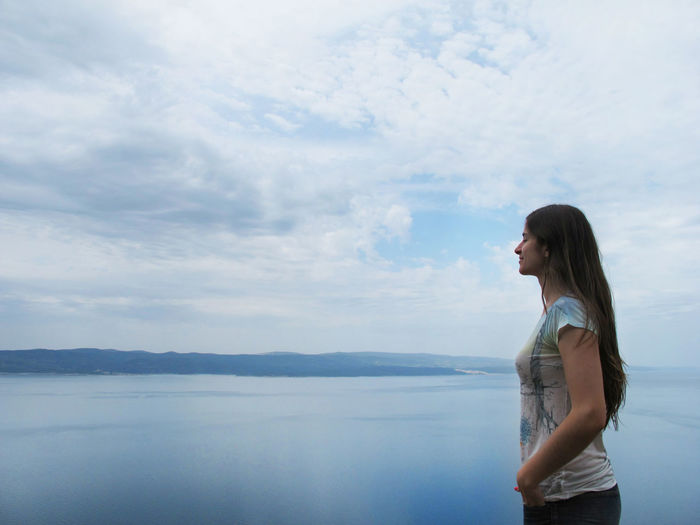 Side view of young woman standing by lake against sky
