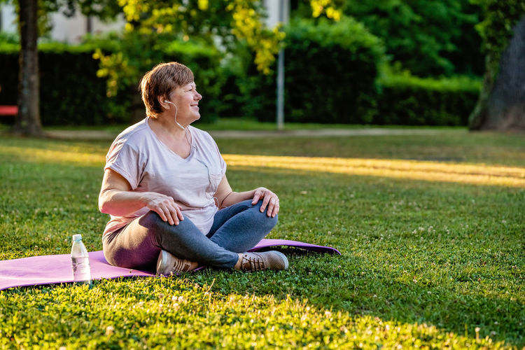 Relaxed senior plus size woman with earphones sitting on yoga mat on in park outdoors resting 