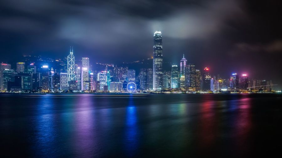 Modern illuminated buildings in front of sea against sky at harbor