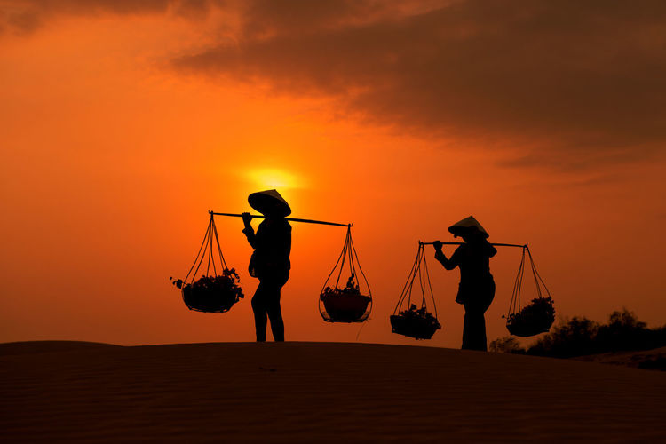Silhouette people carrying plants in baskets against orange sky