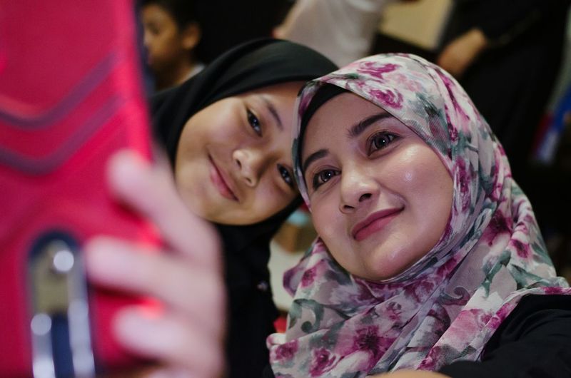 Young woman wearing hijab taking selfie with friend from mobile phone