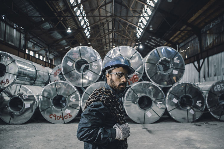Portrait of male worker carrying chain while standing against rolled up steel sheets at factory