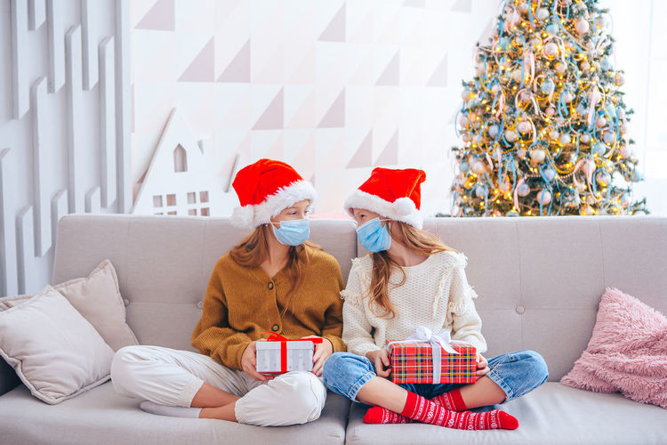 Full length sisters wearing mask sitting on sofa at home