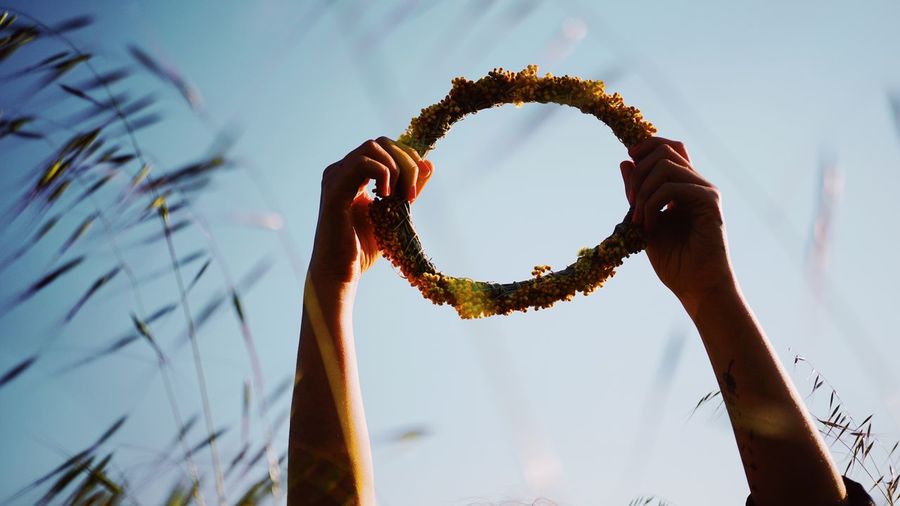 Low angle view of cropped hands holding wreath against sky on field