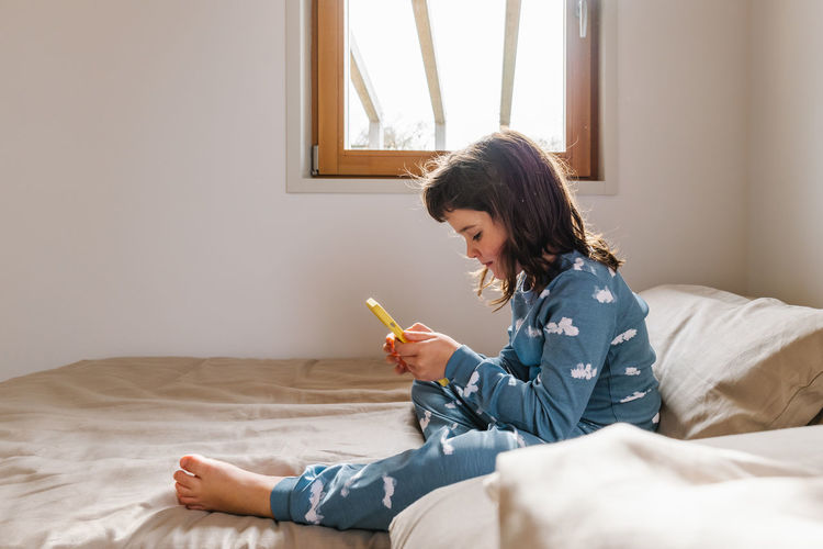 Full body side view of girl in sleepwear playing game on cellphone while sitting on bed near window in light bedroom