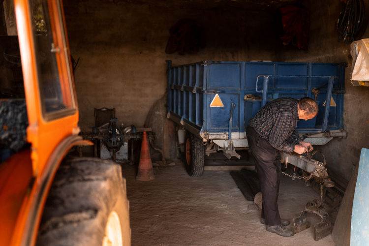 Full body side view of senior male mechanic fixing detail tractor cart while working in rural garage