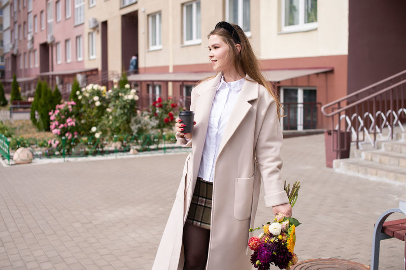 A cute girl in a coat holds a glass of coffee and a bouquet of flowers on the street.