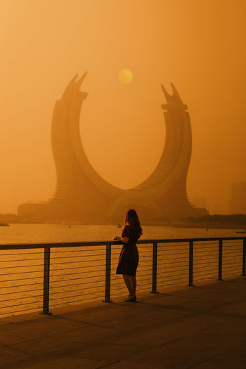Back view of young female tourist with short dark hair in casual clothes standing on embankment and admiring creative moon shaped building against scenic sunset sky in doha