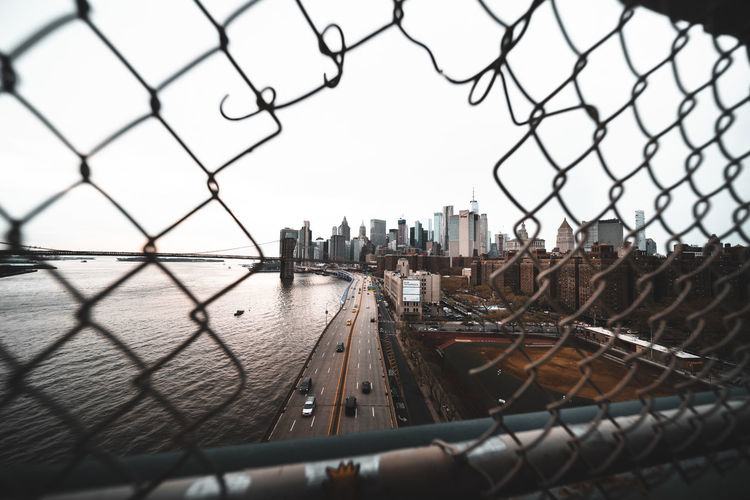 Cityscape by river against clear sky seen through broken chainlink fence