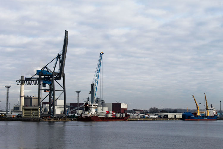 Cranes at commercial dock by river against cloudy sky