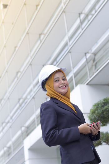 Low angle view of happy female architect holding smart phone while standing against office building