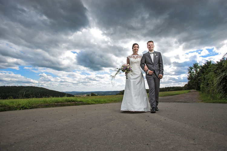 Low angle view of bride and groom walking on road against cloudy sky
