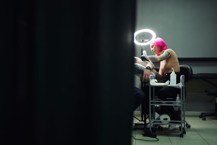 Serious tattoo master with pink hair in gloves using professional tattoo machine while making tattoo on shoulder of client in modern tattoo salon