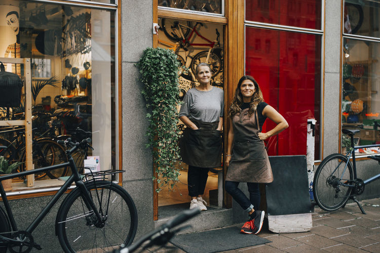 Smiling female coworkers standing at doorway of bicycle shop