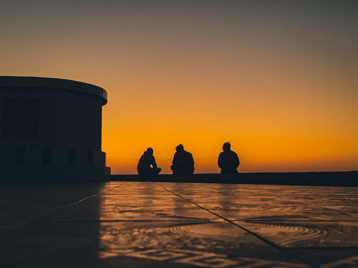 Silhouette people sitting by sea against orange sky