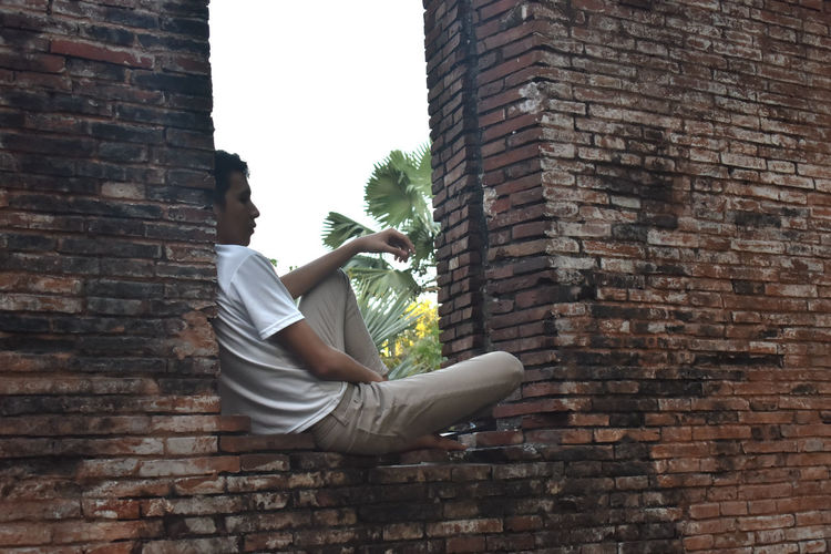 Side view of man sitting on brick wall