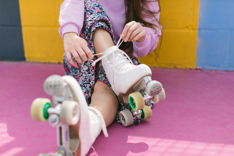 Crop young female in trendy outfit tying shoelaces on white quad roller skates while sitting on colorful playground in sunny day