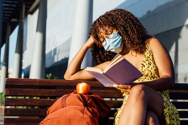 Thoughtful african american female in medical mask sitting on bench in city and enjoying interesting story while resting during coronavirus outbreak