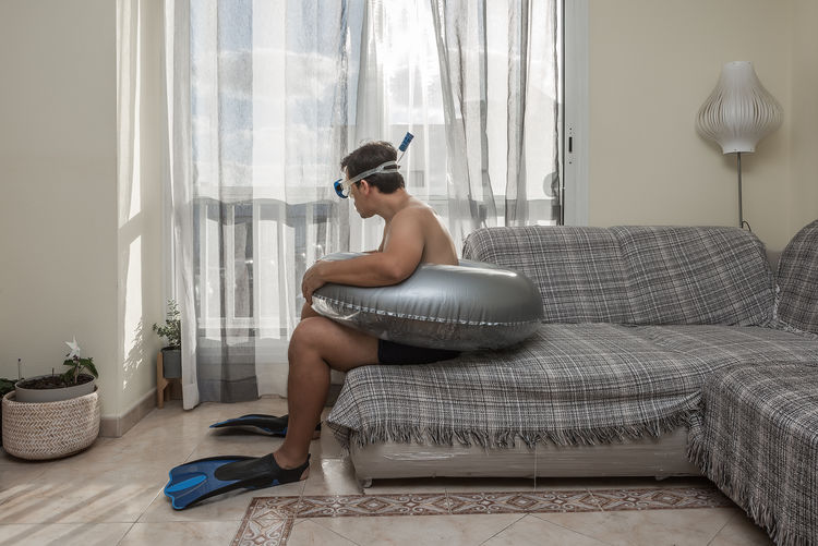 Shirtless man wearing swimming goggles and diving flippers sitting on sofa at home
