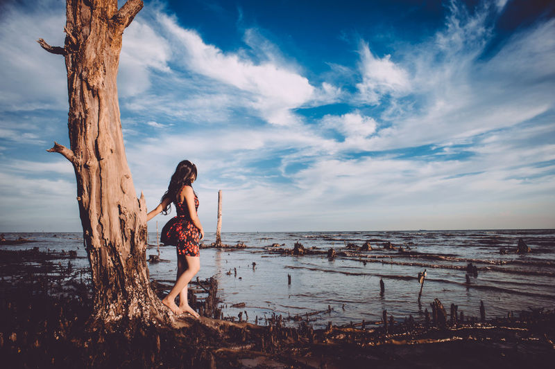 Young woman standing roots of tree at beach against cloudy sky
