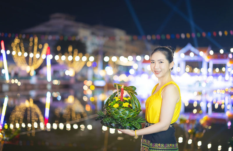 Portrait of young woman standing by illuminated christmas tree at night
