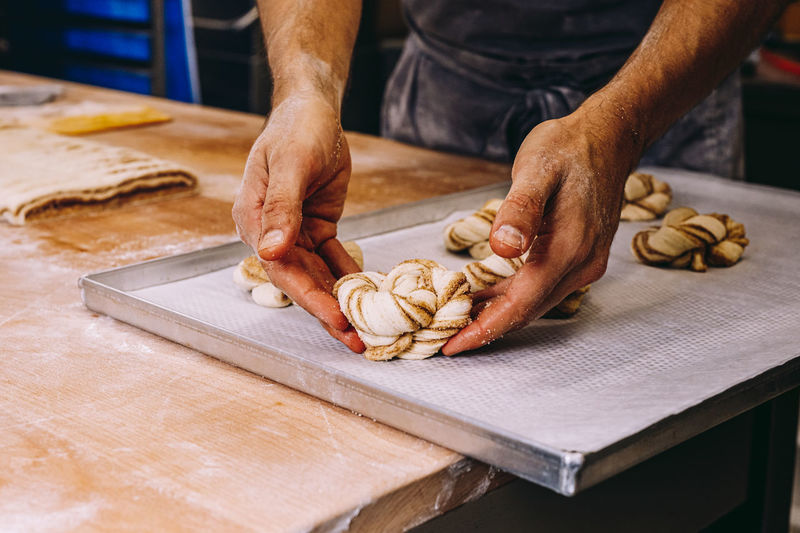 Crop unrecognizable male cook in apron standing at wooden table putting with twisted dough in shape of bun with cinnamon in bakery tray