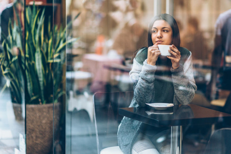 Thoughtful young woman holding coffee cup while looking through window in cafe