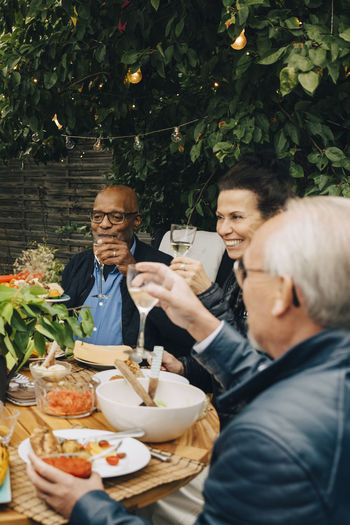Smiling senior woman sitting with male friends while raising toast at dining table during dinner party in back yard