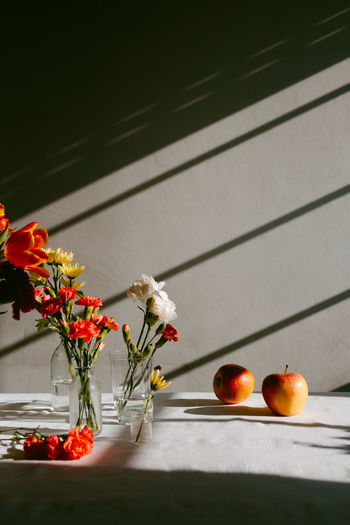 Glass vases with blooming tulips and carnations placed on table near apples with shadow in a wall