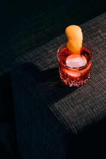 Glass of refreshing negroni cocktail with bitter flavor and ice garnished with orange peel and served on couch arm in dark room
