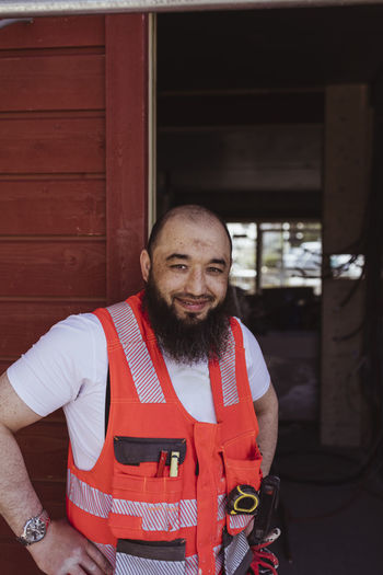 Smiling bearded male construction worker standing with hand on hip at site