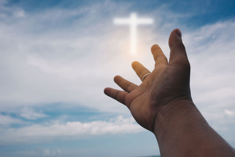 Cropped hand of man gesturing against cross in sky