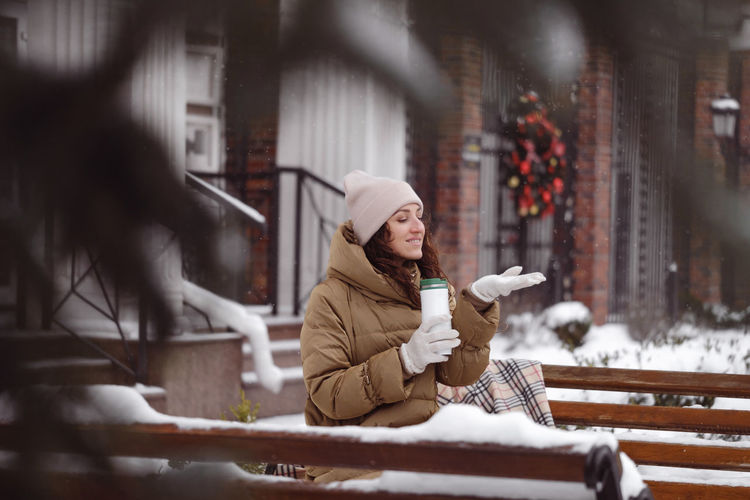 Cheerfully brunette woman drinking from flask outdoors in the city in winter. warming up, enjoying