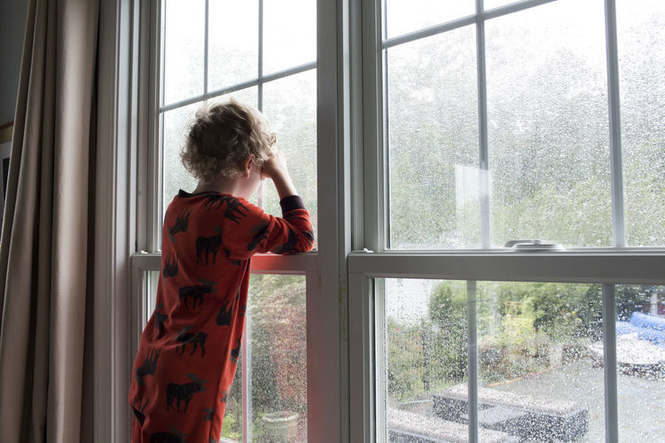 Rear view of young boy with curly hair looking out rain covered window