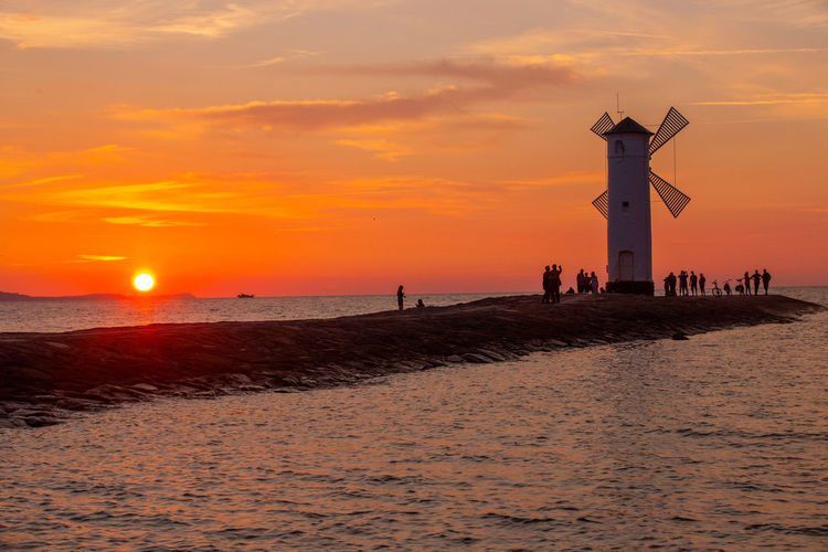 Traditional windmill on pier in sea against orange sky