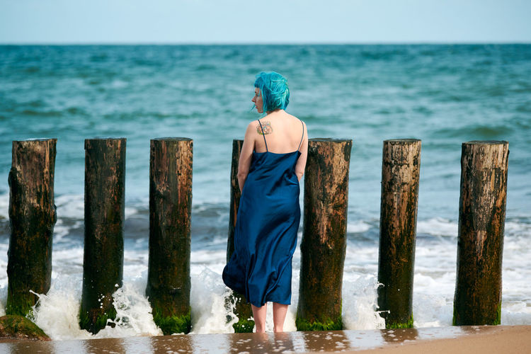 Young blue-haired woman in long dark blue dress standing on sandy beach