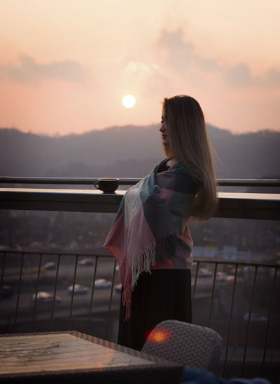 Side view of woman wrapped in shawl standing by railing against sky during sunset