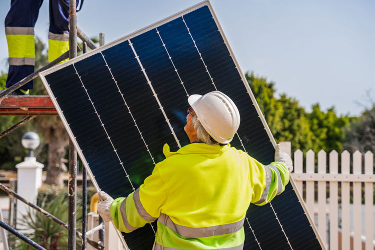 Back view of mature experienced male technician in uniform and helmet standing with solar panel near yellow building while working on installation of renewable energy system