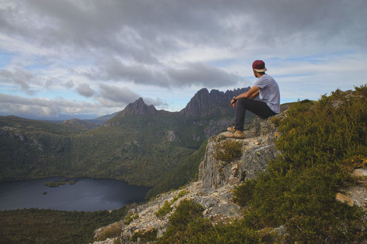 Side view of man sitting on mountain against cloudy sky
