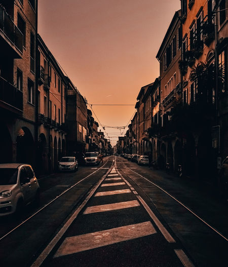 Road by buildings in city against sky during sunset