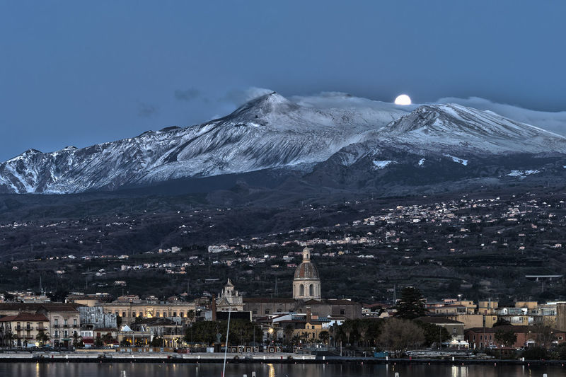 The etna volcano and the full moon at sunset during the blue hour before dawn