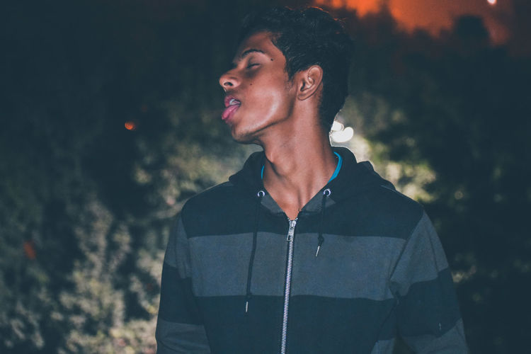 Young man with eyes closed sticking out tongue while standing at night
