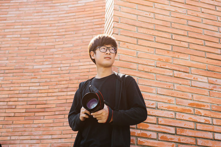 A young asian man stands in front of a brick wall holding a camera and looking forward.